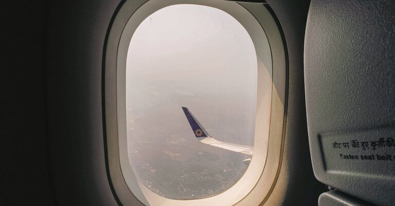 Aerial view from airplane window showing wing over a hazy cityscape, illuminated by daylight.